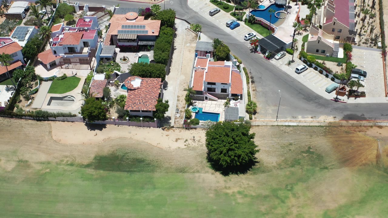 Top-down view of coastal homes, pools, roads, trees, and shallow green water.