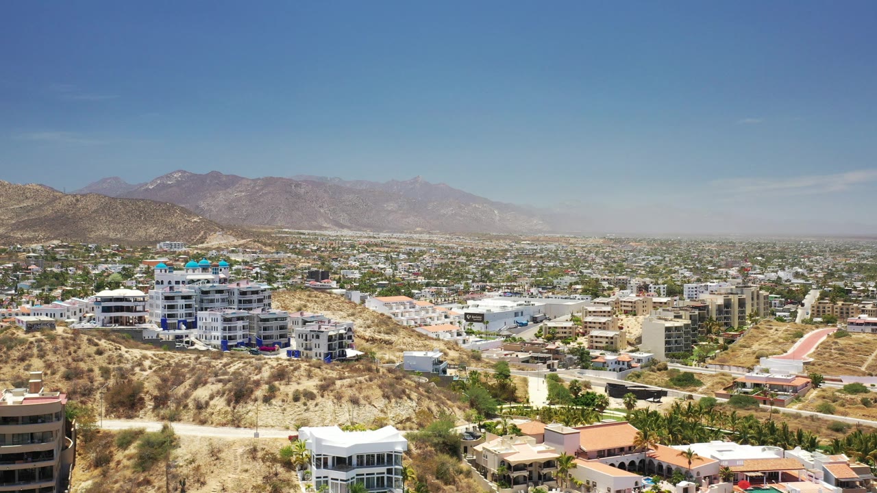 Opening frame of Los Cabos, Mexico, showing dry hillside foreground, white and tan buildings, distant mountains, blue sky, and no visible people.
