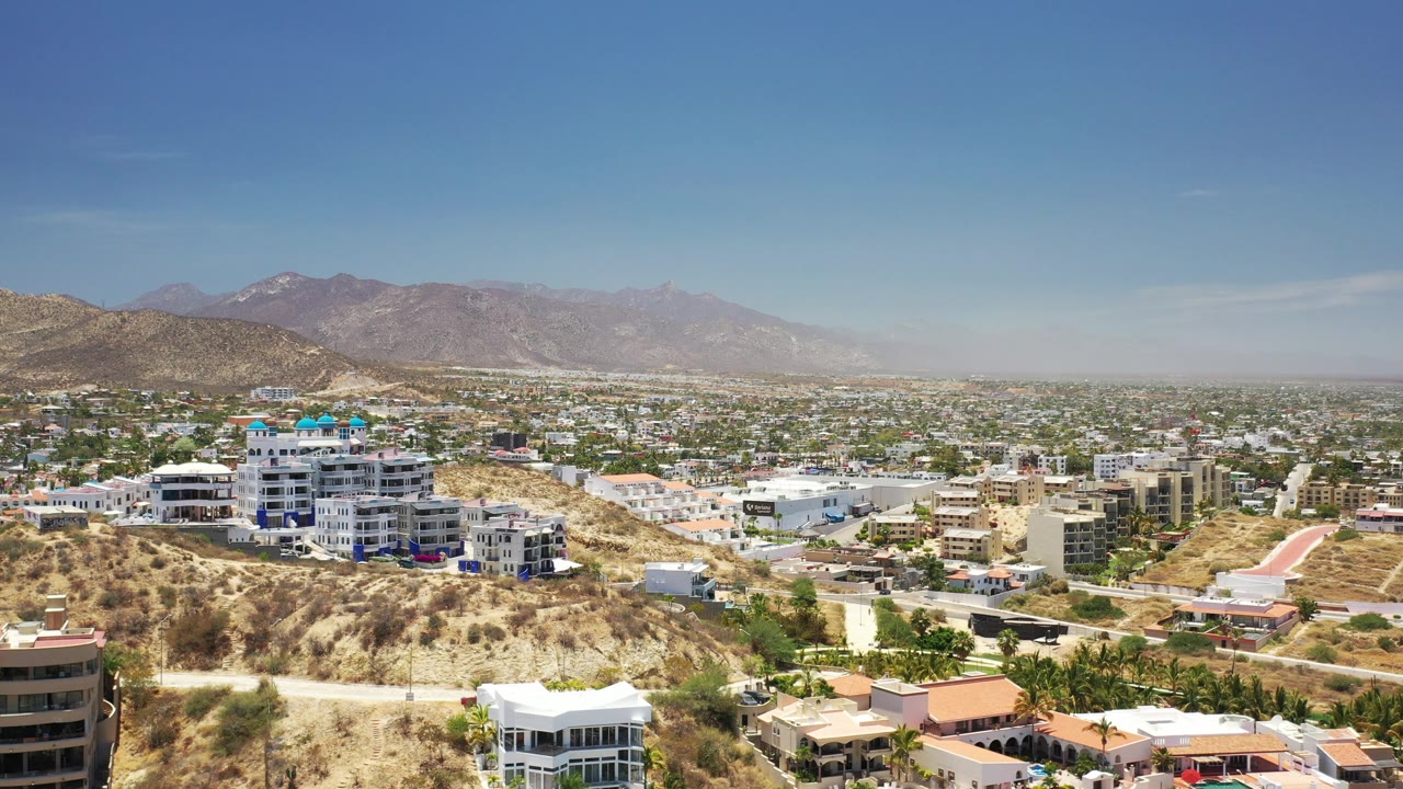 Second frame of Los Cabos, Mexico, with the prominent white hillside building nearer the left, dense town blocks beyond it, mountains, and no visible people.