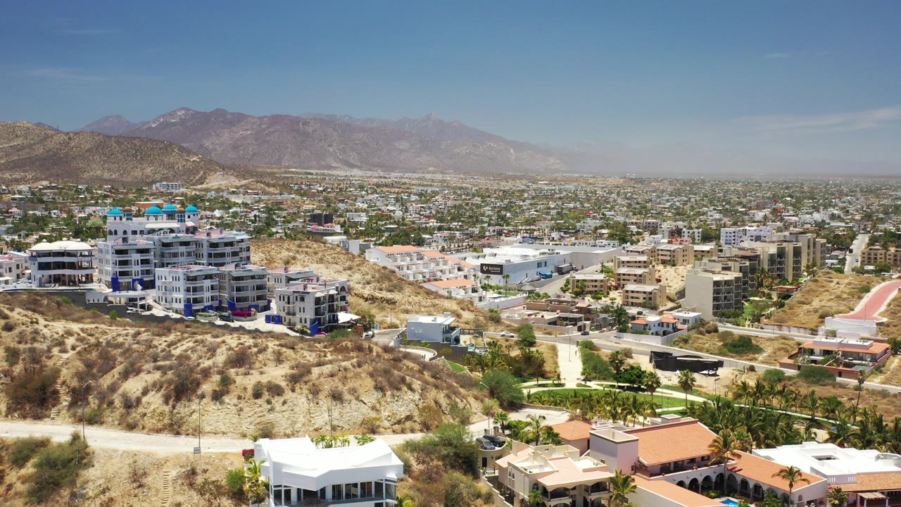 Wide aerial view of hillside homes, city blocks, and mountains under blue sky.