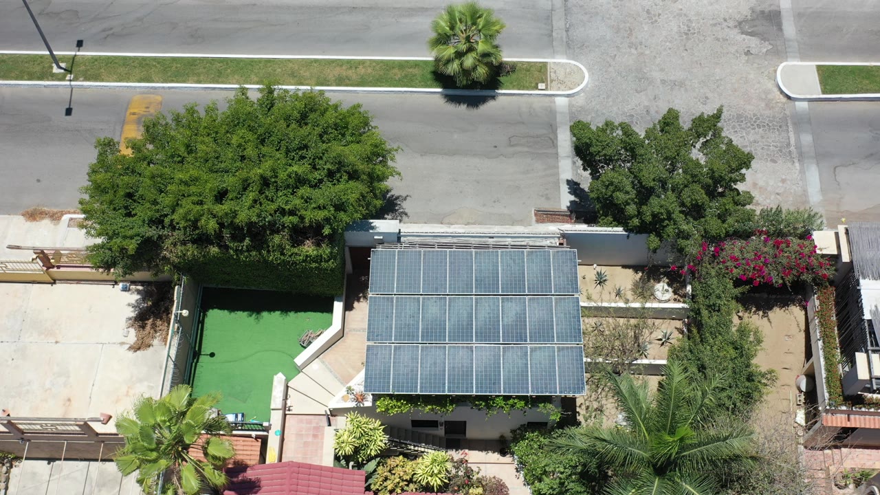 Overhead aerial frame in Los Cabos, Mexico, showing a house with a solar-panel roof, green turf yard, garden walls, palms, and a quiet street, with no visible people.