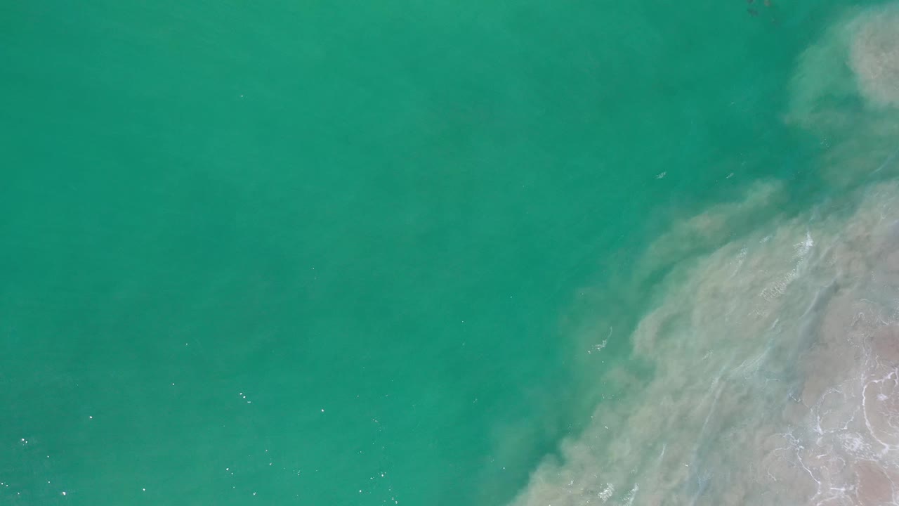 Aerial frame over Los Cabos water showing a cloudy pale sand shape along the right side and open green water across the left, with no visible people.