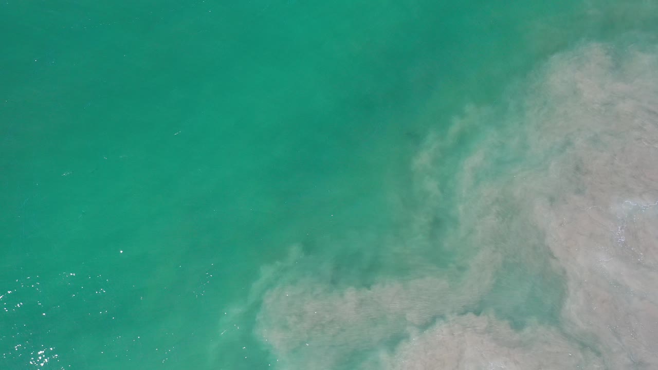 Overhead aerial view of turquoise water in Los Cabos, Mexico, with pale sandy surf patterns entering from the right and no visible people.