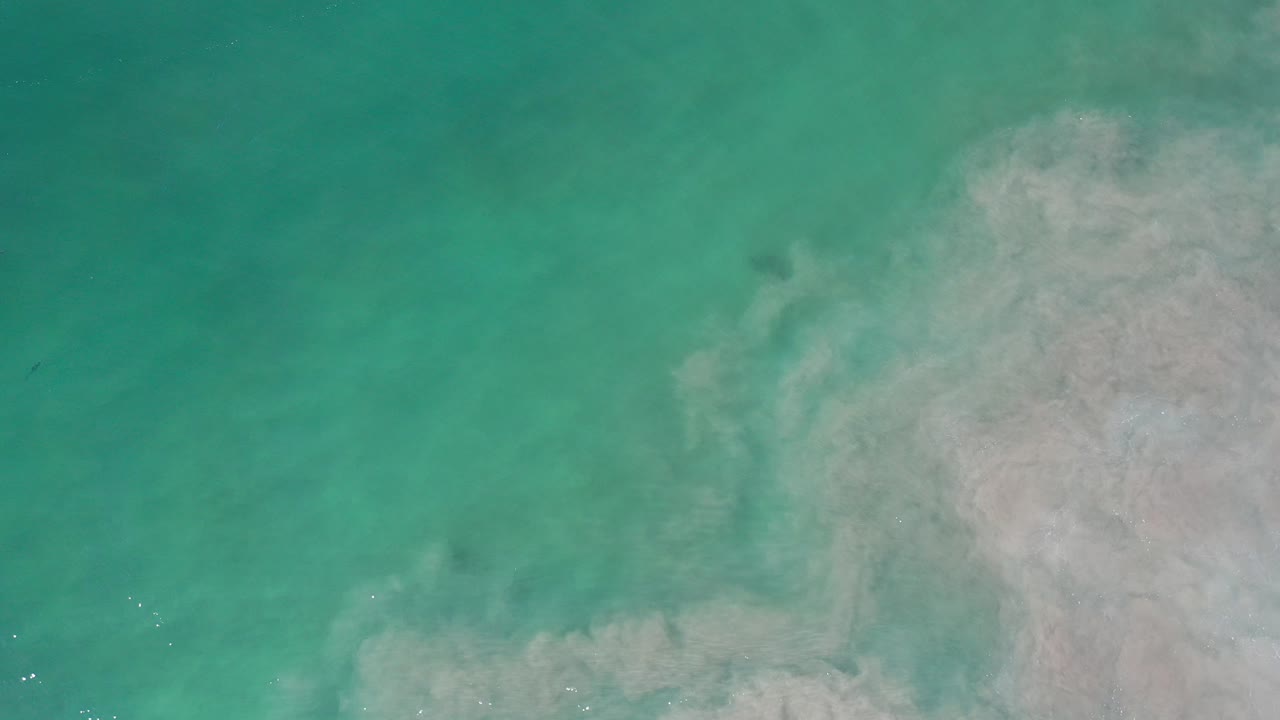 Overhead view of turquoise sea in Los Cabos, Mexico, with a pale cloudy sand or foam area entering from the right and no visible people.