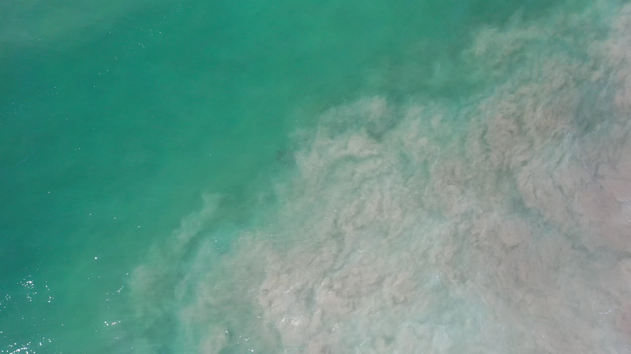 Top-down view of turquoise water and a pale sand cloud spreading through shallow water in Los Cabos, Mexico, with no visible people.