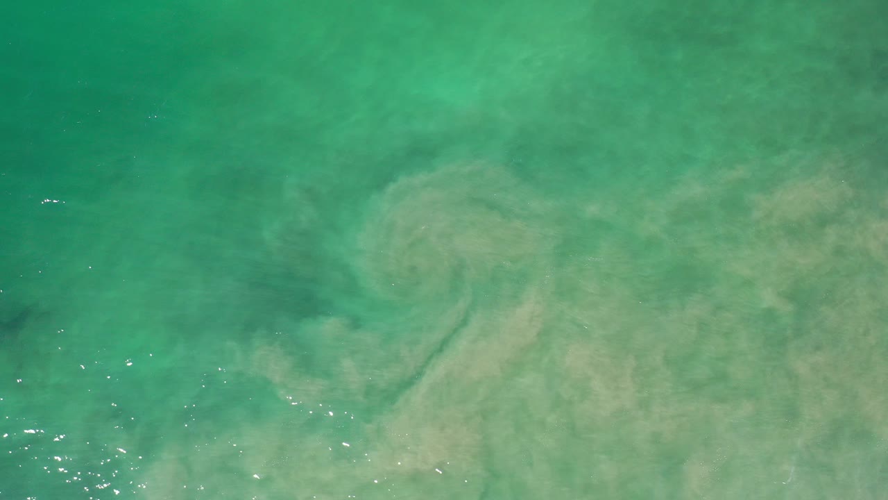 Overhead view of turquoise-green Los Cabos water with a broad pale brown sediment cloud under the surface and no visible people.