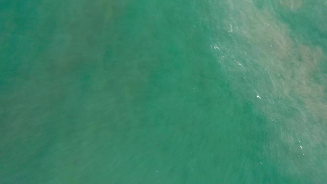 Aerial frame over Los Cabos ocean water, showing faint sandy shapes under the green surface and no visible people.