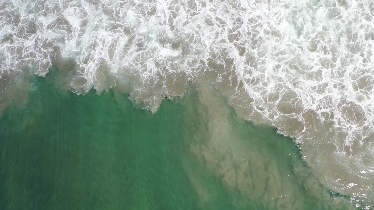 Top-down view of foamy wave edges and translucent green shallows over a sandy seabed in Los Cabos, Mexico, with no visible people.