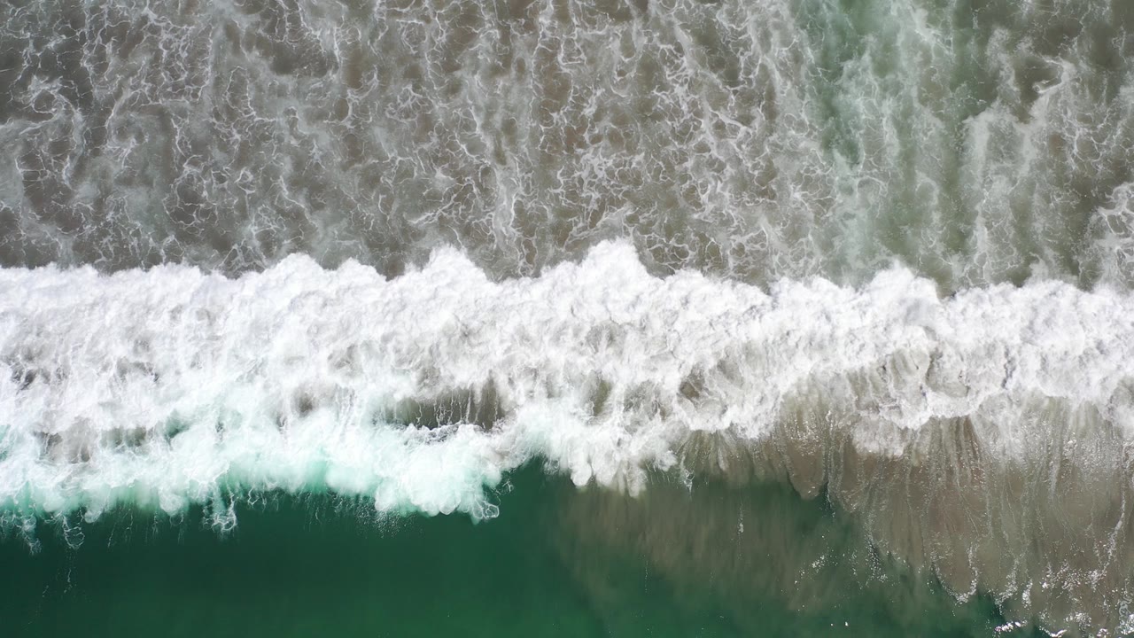 Overhead view of Los Cabos ocean water with a white breaking wave stretching across green and gray shallows, no visible people.