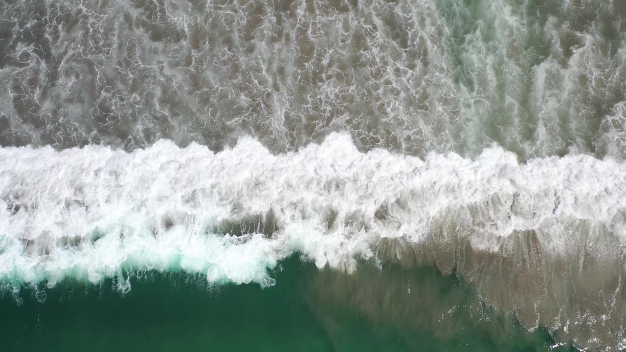 Aerial Los Cabos frame showing white foam spreading in a horizontal strip over muted green ocean and darker shallow water, no visible people.