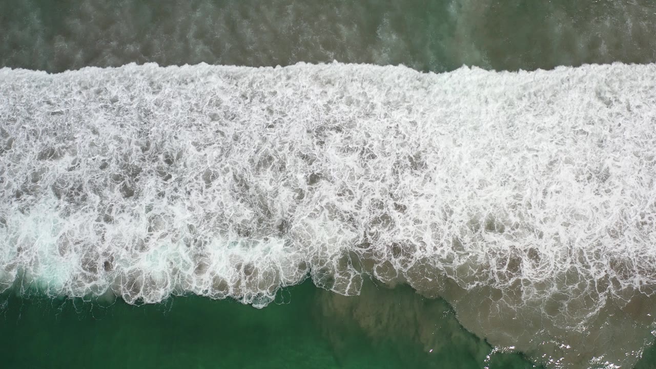 Overhead view in Los Cabos, Mexico, showing a broad white surf band over green water and a narrow tan beach edge, with no visible people.