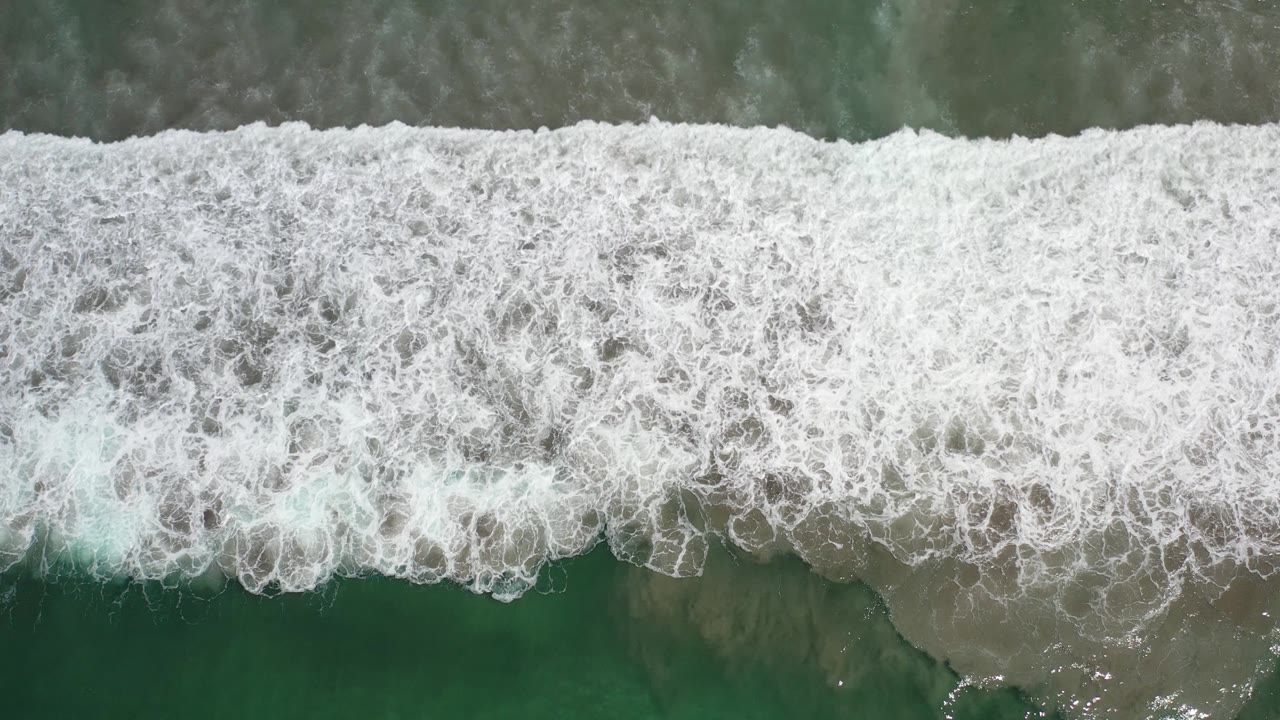 Overhead Los Cabos beach frame with dense white foam rolling across the center and darker green water visible above and below, with no visible people.