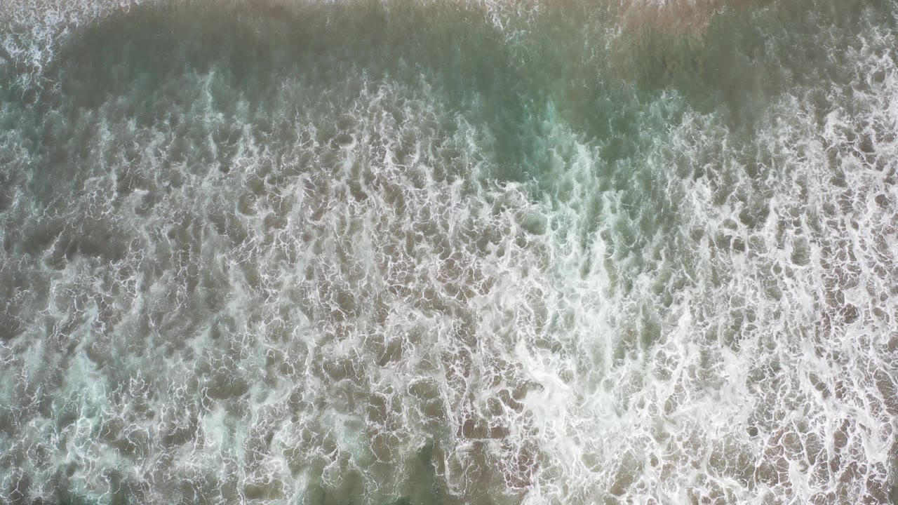 Top-down view of foamy green surf near pale sand in Los Cabos, Mexico, with no visible people.