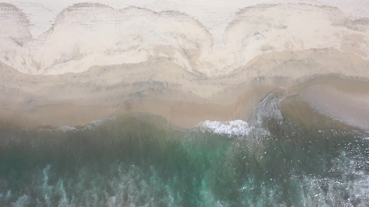 Aerial shoreline view in Los Cabos, Mexico, with a curved band of foam moving across wet sand and no visible people.