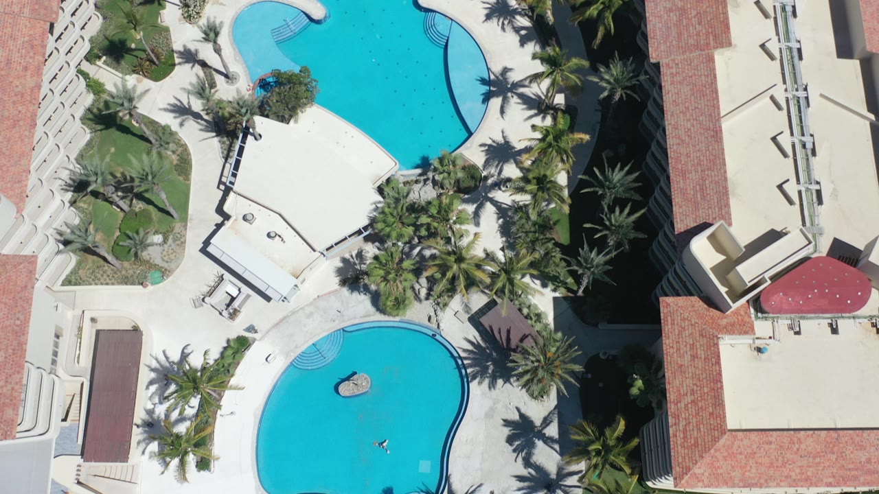 Overhead view of two resort pools beside palm trees and hotel roofs.