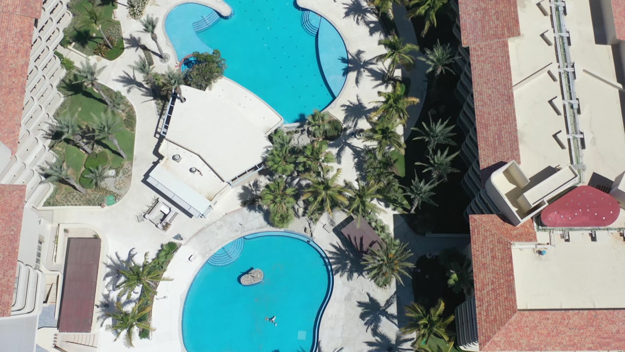 Overhead Los Cabos resort view with the upper pool, lower pool, palm shadows, white walkways, and a hotel wing along the right edge, with no visible people.