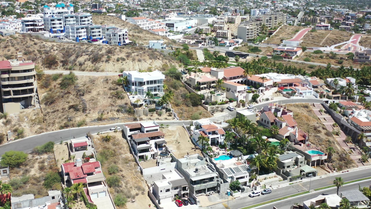 Opening aerial frame over a Los Cabos hillside community with white homes, red tile roofs, paved roads, pools, dry terrain, and no visible people.