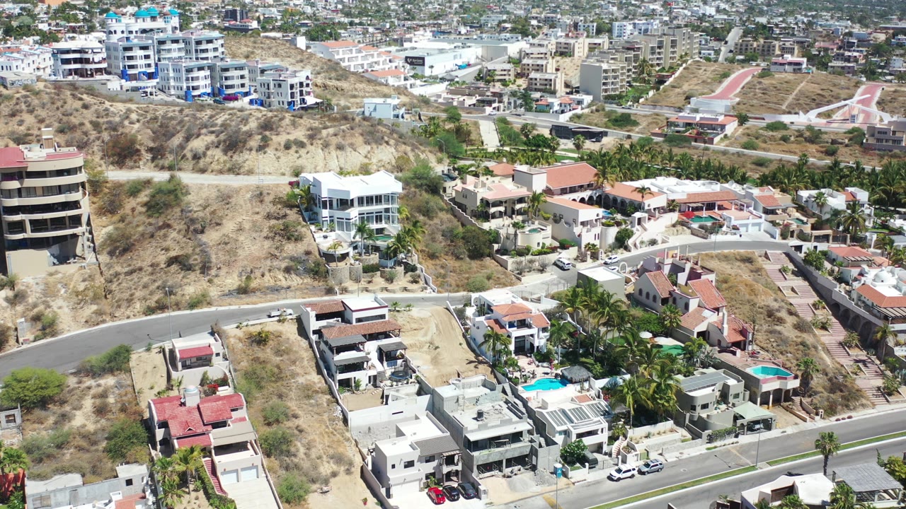 Aerial frame in Los Cabos showing the hillside neighborhood from slightly farther along the pass, with a curved road, pale homes, pools, palms, and no visible people.
