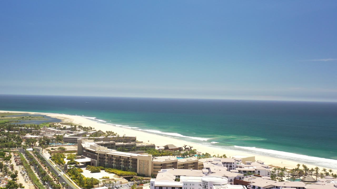 Aerial view over Los Cabos beachfront hotels, pale sand, turquoise shoreline, and deep blue ocean under clear sky, with no visible people.