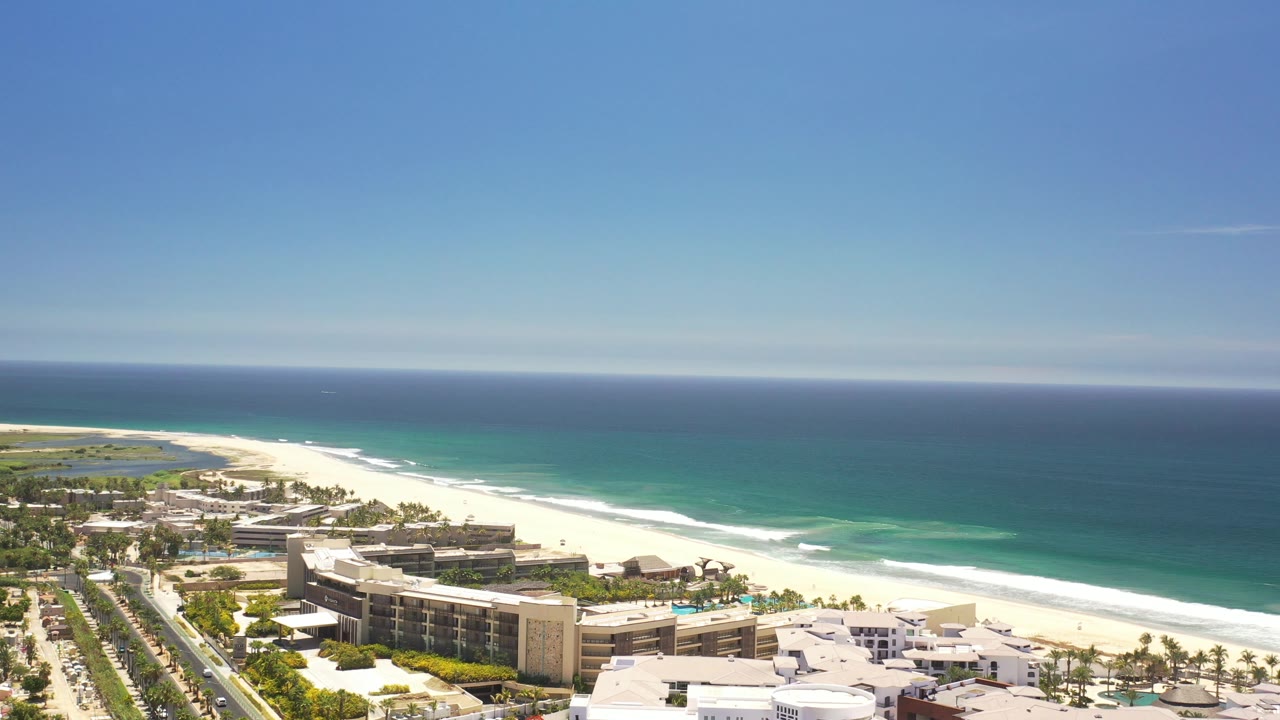 Wide aerial of coastal hotels, palm landscaping, beach, and open water.