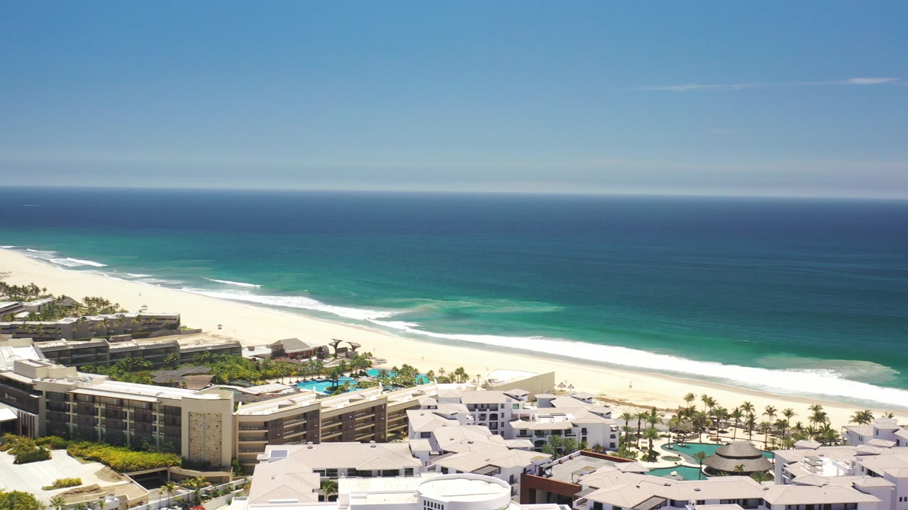 Aerial view of beachfront buildings, pools, sand, surf, and blue ocean.