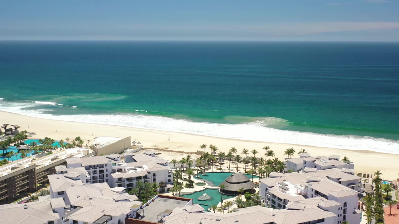 Elevated Los Cabos resort view with a curved swimming pool near the lower center, white resort buildings, rows of palms, light sand, breaking surf, and deep blue ocean.
