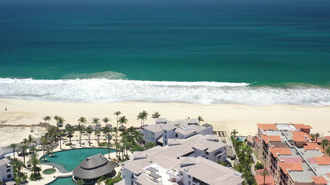 Aerial view of ocean surf, empty beach, resort pools, palms, and rooftops.