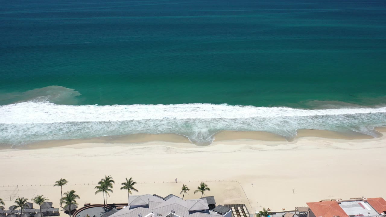 Aerial beach view with green ocean, white surf, pale sand, palms, and rooftops.