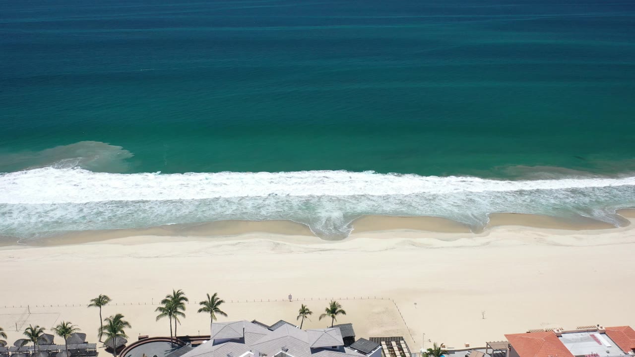 Aerial view over Los Cabos shoreline as foamy surf spreads across pale sand, with green sea above, rooftops near the lower border, and no visible people.
