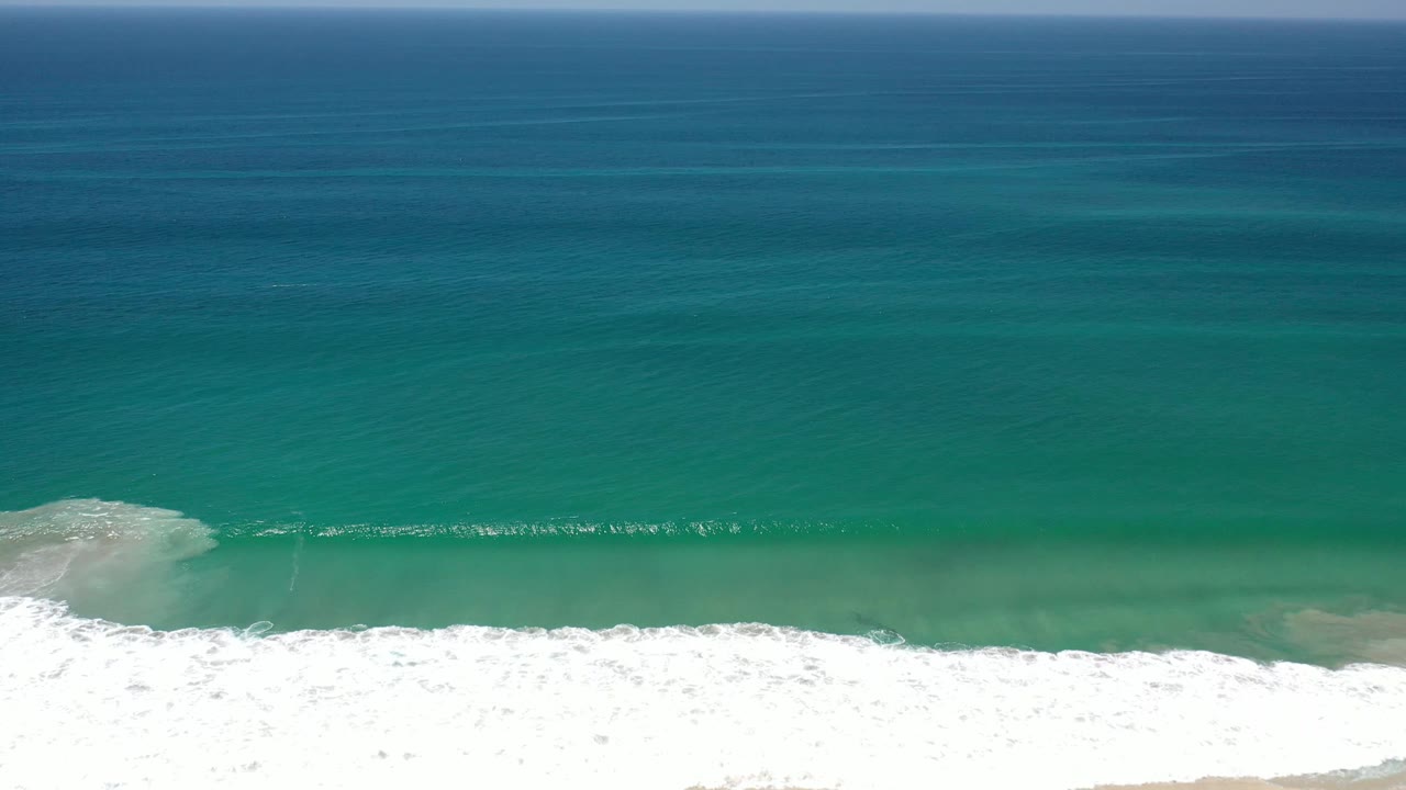 Wide Los Cabos ocean frame with a small white wave line crossing the shallows, darker blue water beyond, and no visible people.