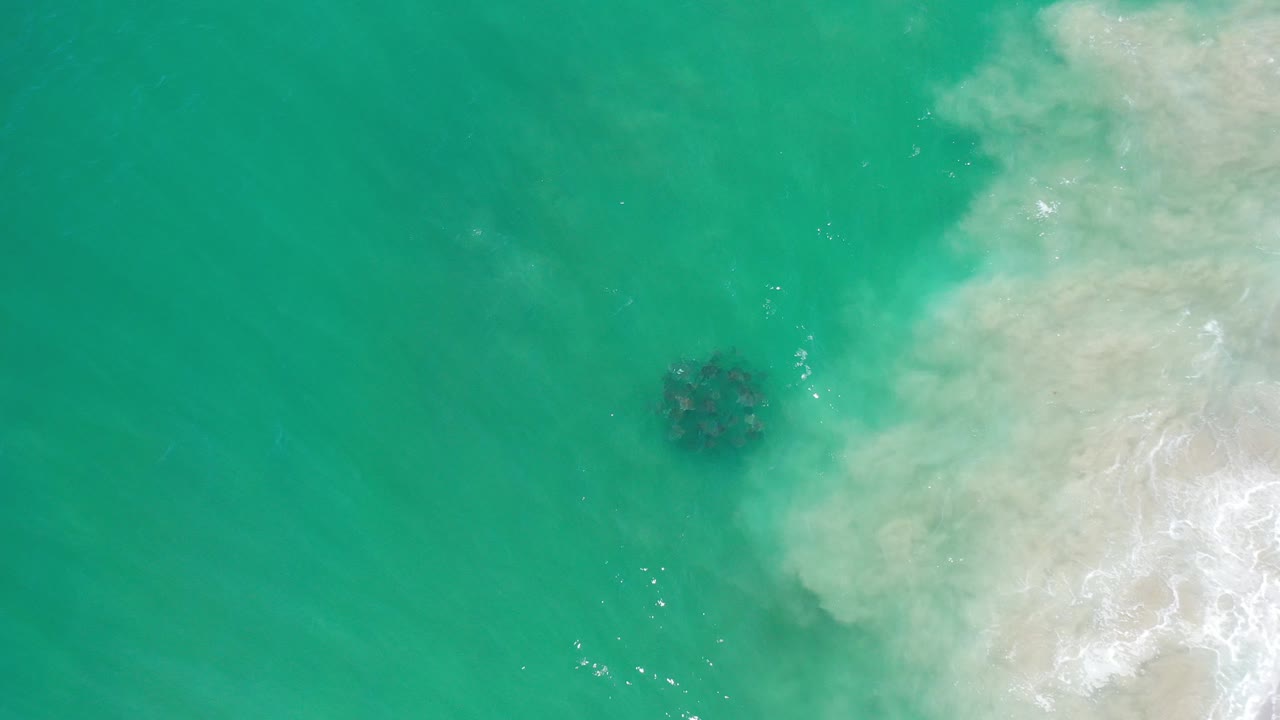 Overhead view of turquoise water in Los Cabos, Mexico, with pale foamy surf entering from the right and a dark underwater patch near the center, no visible people.