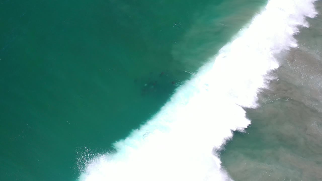 Aerial frame in Los Cabos, Mexico, with dense white foam pushing diagonally across green ocean water and gray shallow water at the edge, with no visible people.