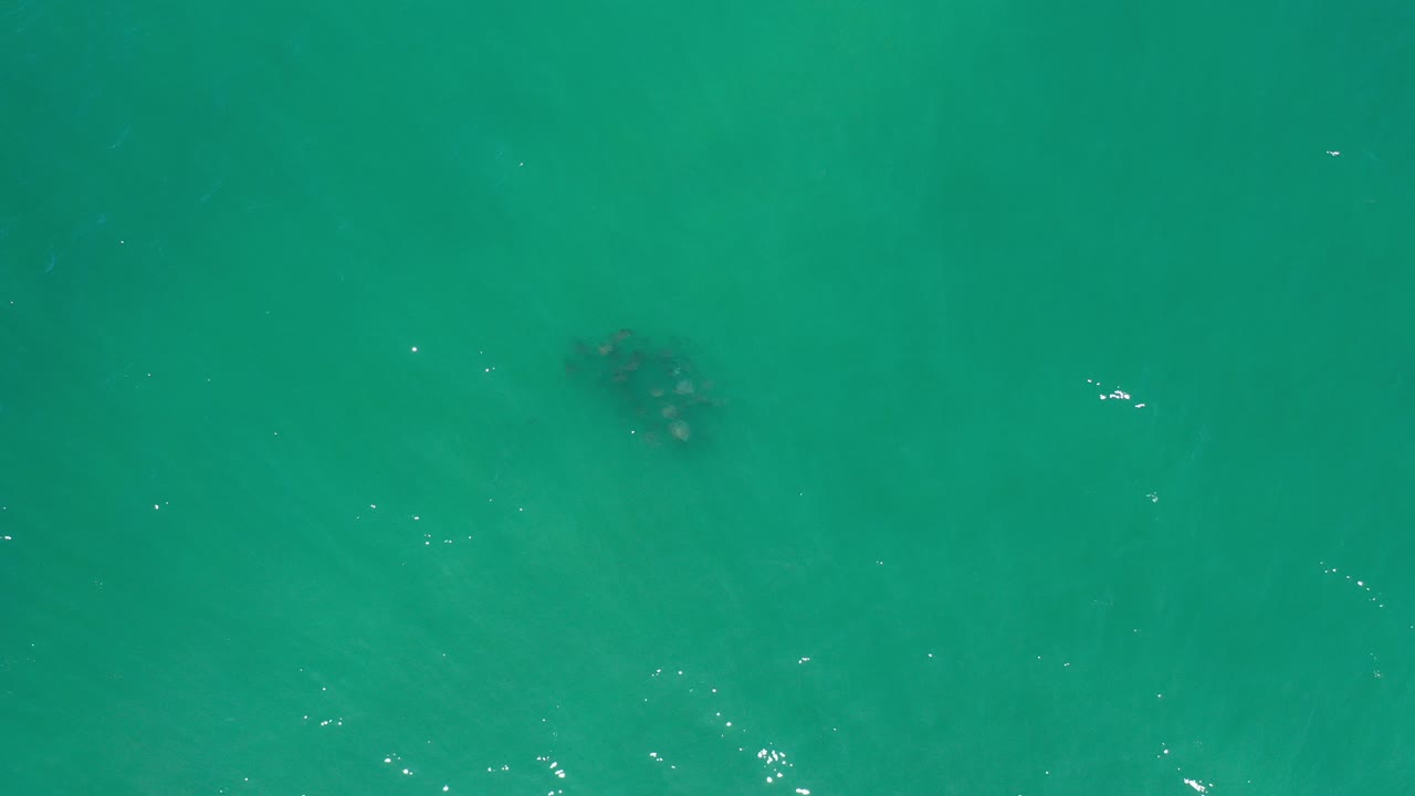 Top-down view of turquoise ocean in Los Cabos, Mexico with scattered sun reflections, a small dark underwater patch near the upper left, and no visible people.