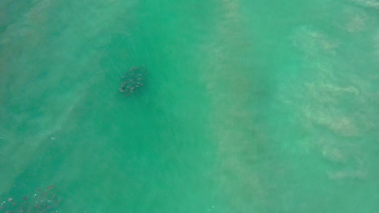 Top-down turquoise water with faint underwater sand shapes and a dark patch.