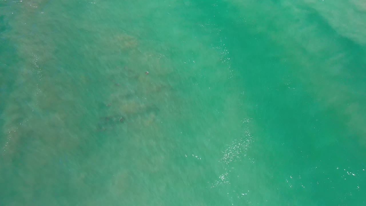 Aerial Los Cabos water frame showing faint seabed shapes under green-blue water, with a few bright reflections and no visible people.