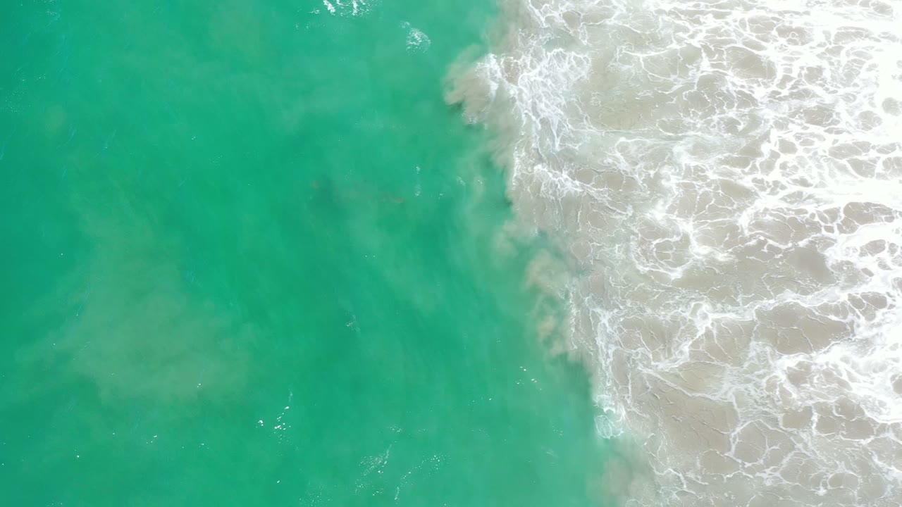 Top-down view of bluish green water and white surf meeting pale sand in Los Cabos, Mexico, with no visible people.