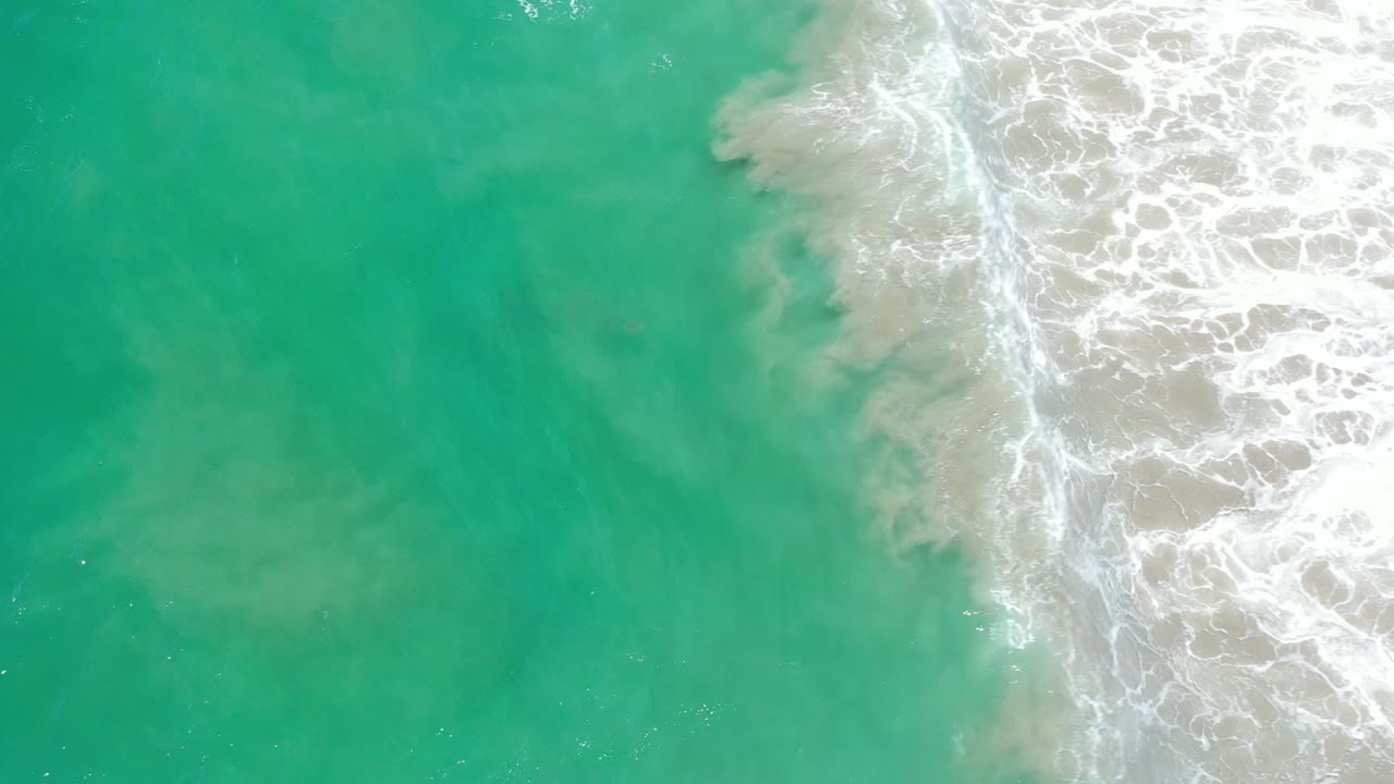 Aerial-style frame of green-blue Los Cabos water as a foamy wave edge pushes diagonally from the right, with no visible people.