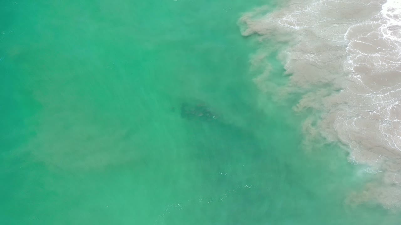 Aerial-looking frame over green-blue water in Los Cabos, Mexico, where a faint dark fish group appears below the surface near the right-side surf, with no visible people.