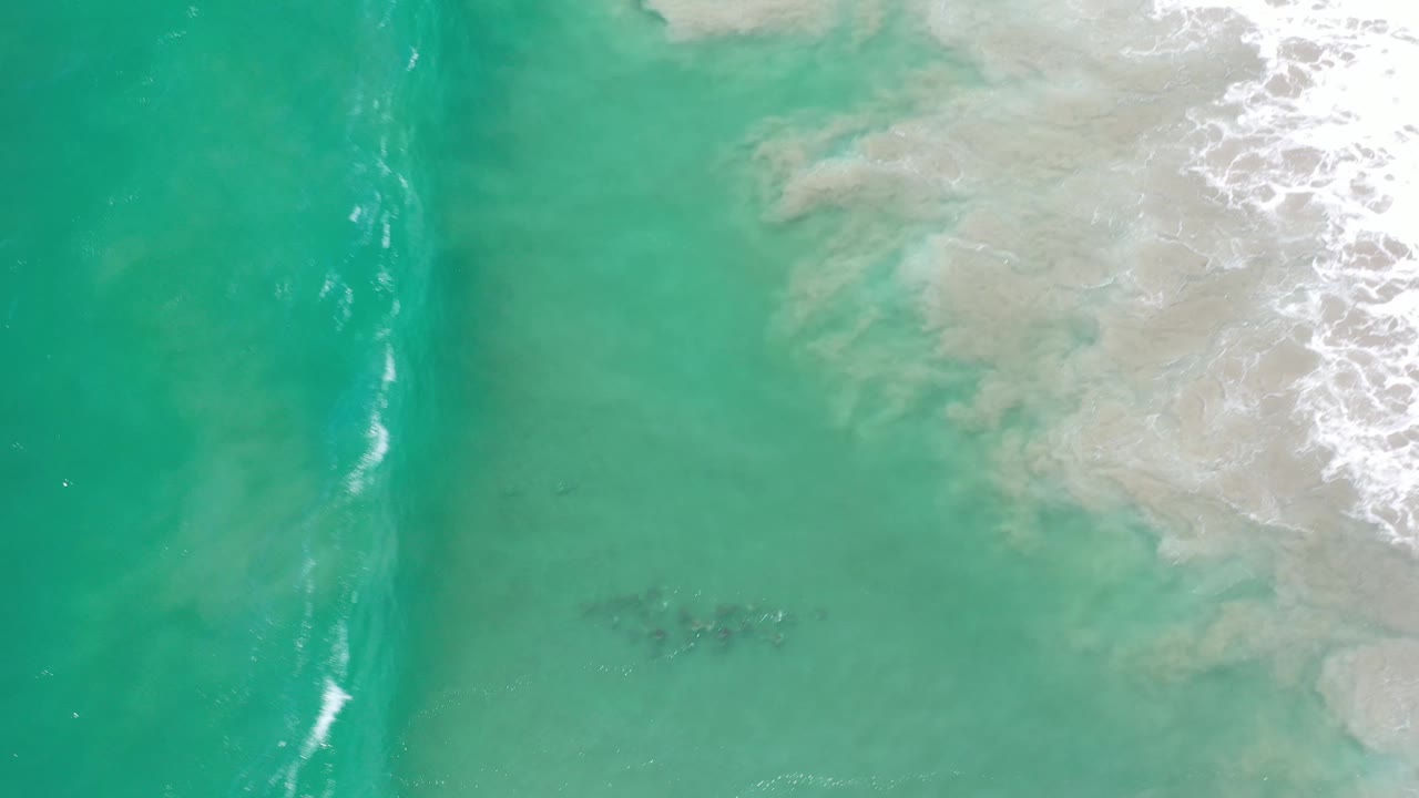 Aerial Los Cabos shoreline frame with green water, faint underwater patches, and white foam gathering near the upper right, with no visible people.