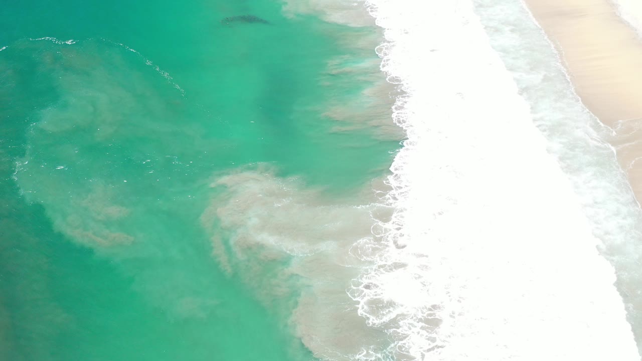 Overhead view of turquoise water and white surf reaching a pale sandy beach in Los Cabos, Mexico, with no visible people.