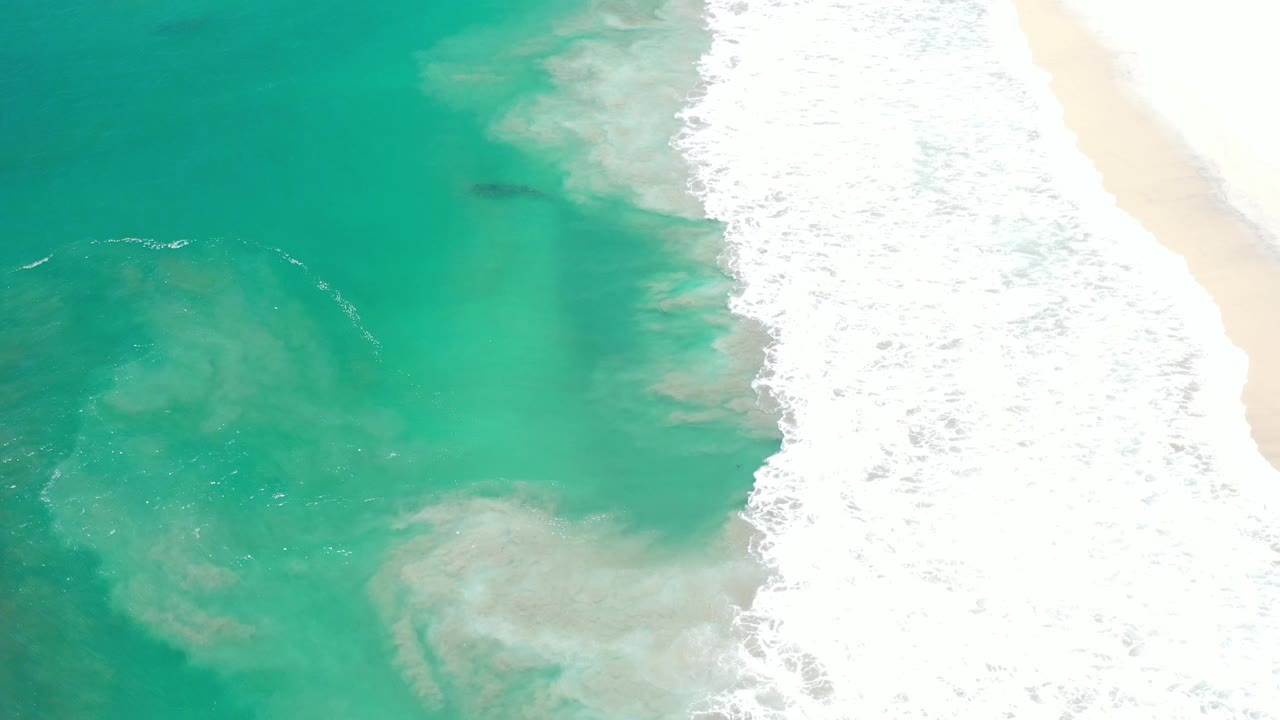 Aerial Los Cabos beach frame with a dense white foam band along the right, green-blue shallow water across the center, pale sand at the edge, and no visible people.