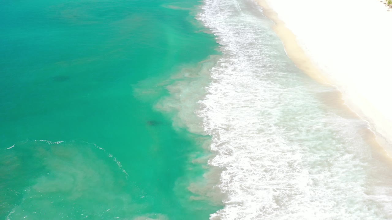 High aerial view over Los Cabos, Mexico, with turquoise water, a diagonal line of white surf, pale sand at the right edge, and no visible people.
