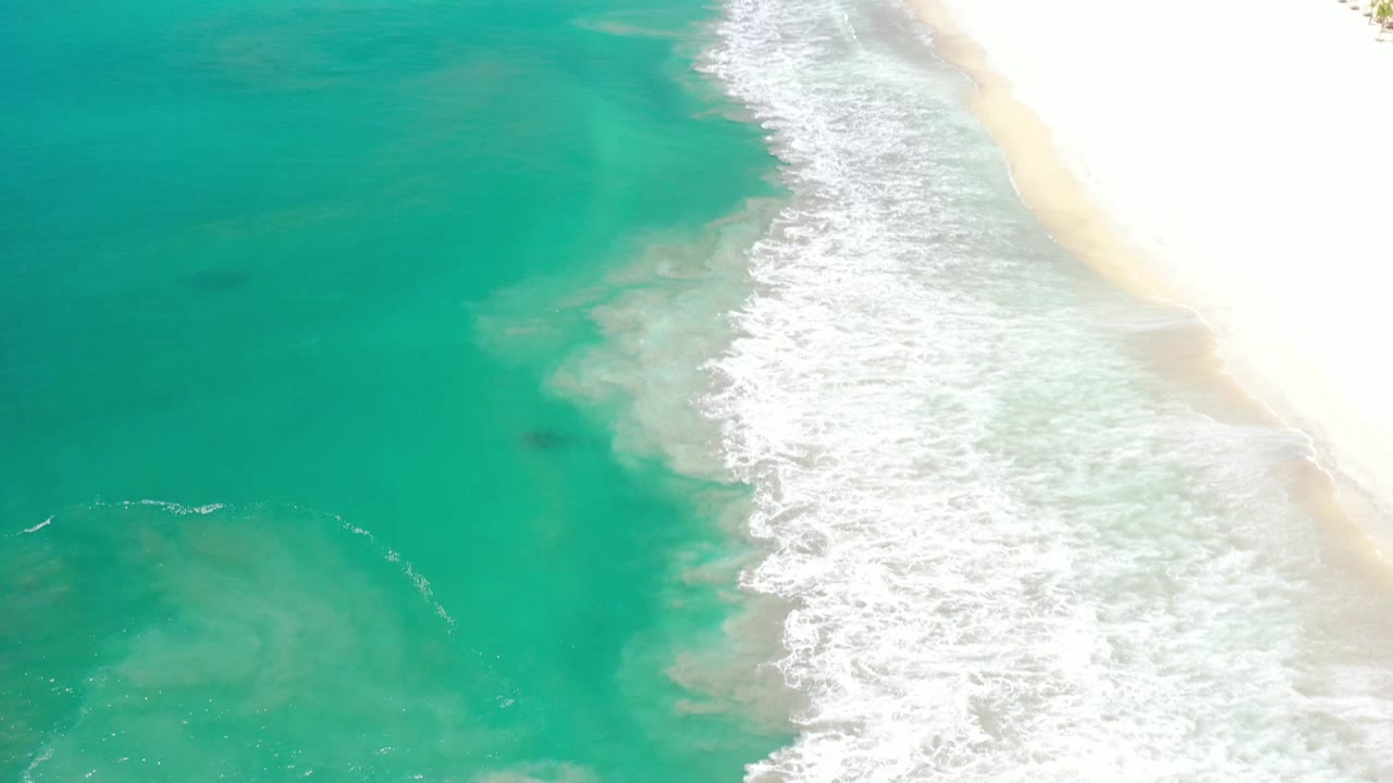 Los Cabos shoreline seen from above, with green-blue ocean on the left, foamy surf crossing the center, bright sand on the right, and no visible people.