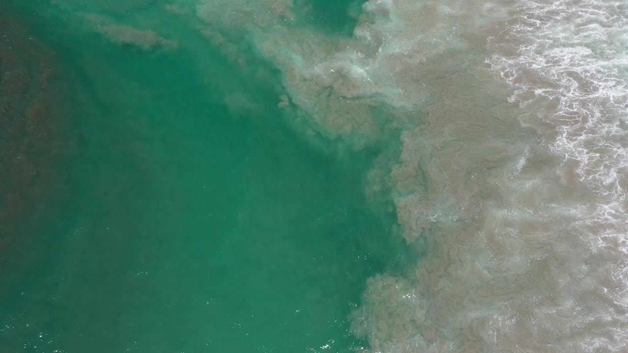 Overhead view in Los Cabos, Mexico of turquoise-green water meeting white surf and suspended sand, with no visible people.