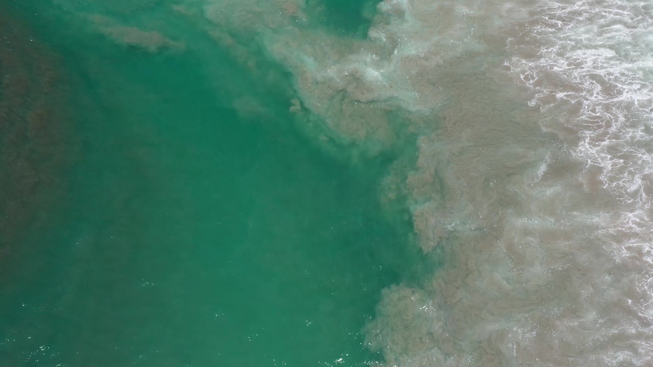 Overhead Los Cabos ocean frame with a large green water area on the left and a pale foamy break pushing through sand on the right, no visible people.