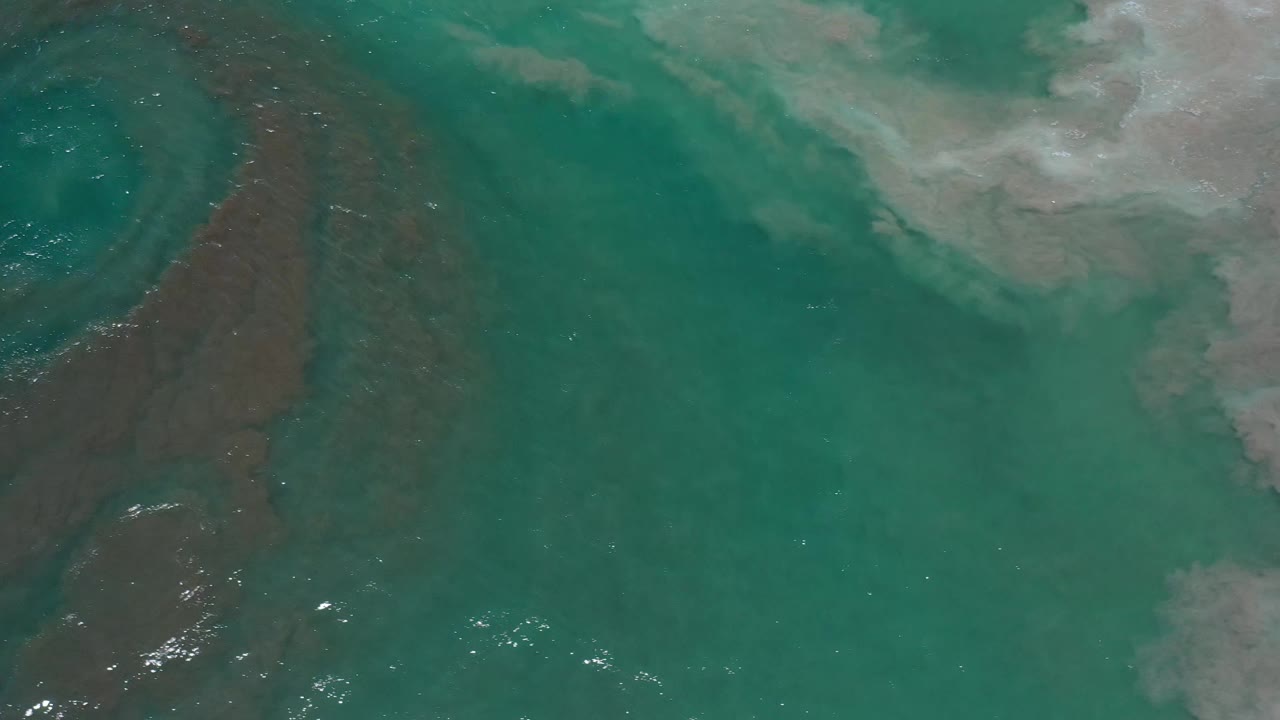 Overhead Los Cabos ocean frame with green-blue water, a faint brown current band on the left, white foam across the upper right, and no visible people.