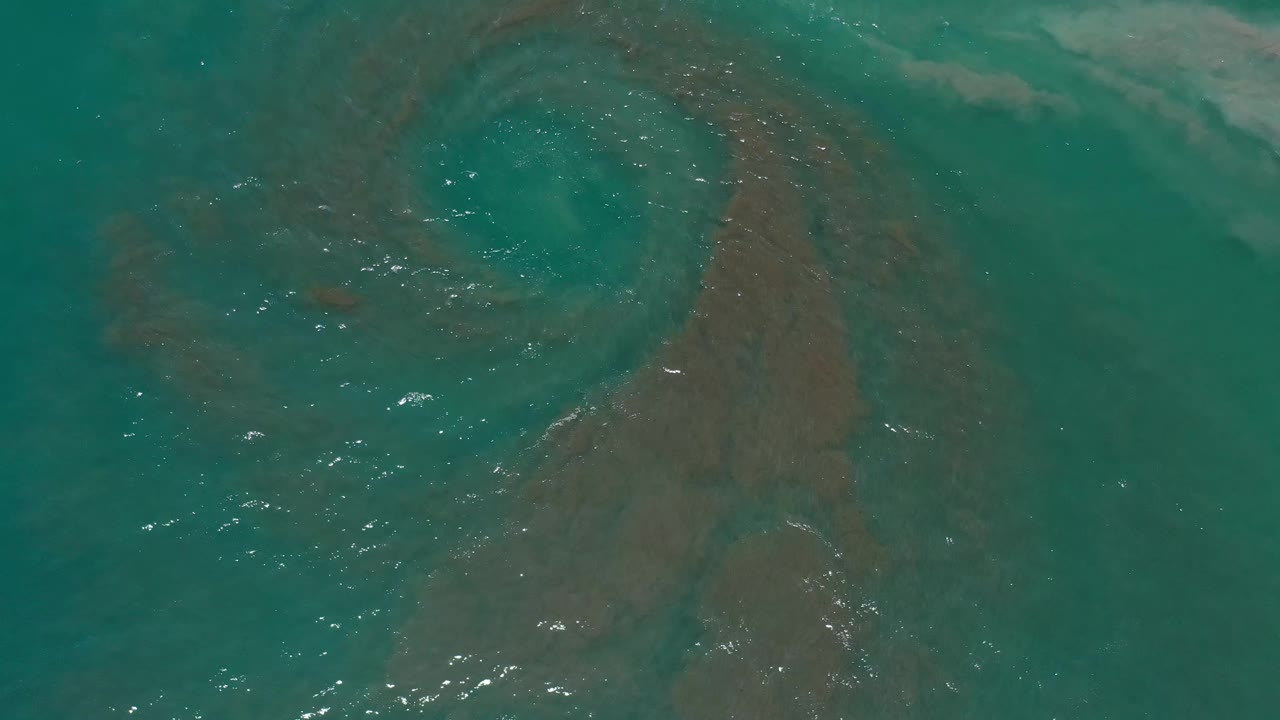 Blue-green water in Los Cabos, Mexico shows a curved brown underwater pattern near center with bright ripple highlights and no visible people.