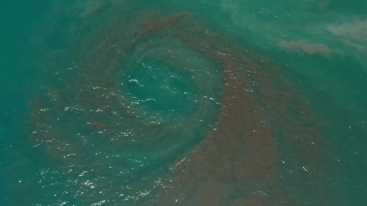 Overhead Los Cabos water frame with small waves crossing a brownish circular underwater feature and scattered highlights on the green surface, with no visible people.