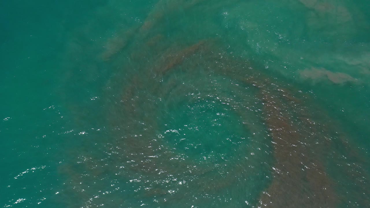 Top-down view of teal sea water in Los Cabos, Mexico, with brown tide bands below the rippled surface and no visible people.