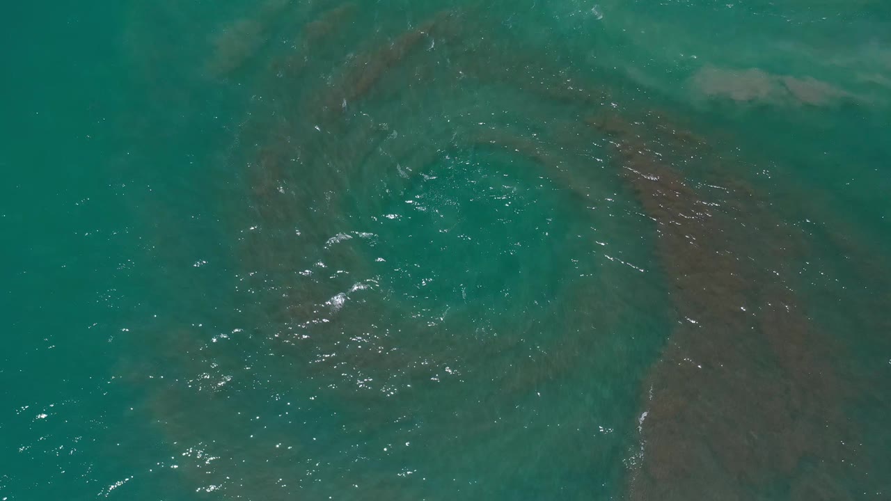 Turquoise water in Los Cabos shows curved brown sand bands below the surface with scattered bright glints and no visible people.